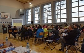 Sala gimnastyczna w Mszanie. Widok od tyłu na siedzące na krzesłach kilkadziesiąt os&oacute;b. Na wprost widoczny ekran z niebiesko-ż&oacute;łtą ukraińską flagą