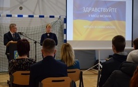 Sala gimnastyczna w Mszanie. Widok od tyłu na siedzące na krzesłach kilkadziesiąt os&oacute;b. Na wprost widoczny ekran z niebiesko-ż&oacute;łtą ukraińską flagą i stojące za m&oacute;wnicą 2 osoby 